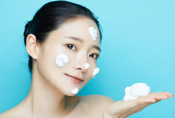 DODECANE – quartz-chem Woman applying skincare cream on face with several dollops of white cream on her cheek and forehead, holding a blob of foam on her hand against a blue background.